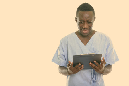 Studio shot of young sad black African man patient reading on clipboard while looking shocked