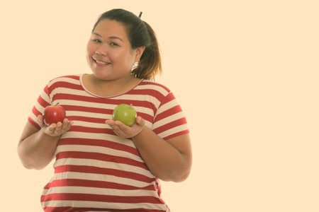 Studio shot of young happy fat Asian woman smiling while holding red apple and green appleの写真素材