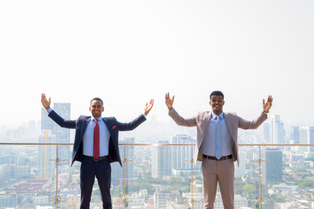 Portrait of two African businessman with arms raised at rooftop with arms raised against city viewの写真素材