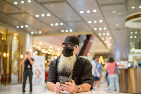 Man wearing protective surgical mask with sunglasses and cap for safety against corona virus while thinking and using mobile phoneの写真素材