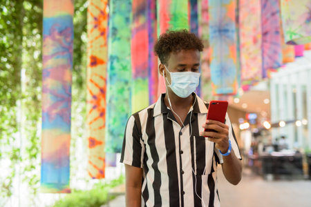 Portrait of handsome black African man wearing face mask in shopping mall while using mobile phoneの写真素材
