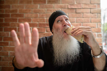 Hungry man with long gray beard eating taco while getting disturbed and showing stop gestureの写真素材