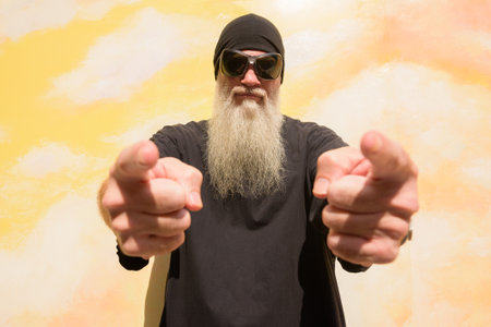 Summer portrait of mature man with long gray beard wearing sunglasses against cloudy background wall while pointing fingerの写真素材