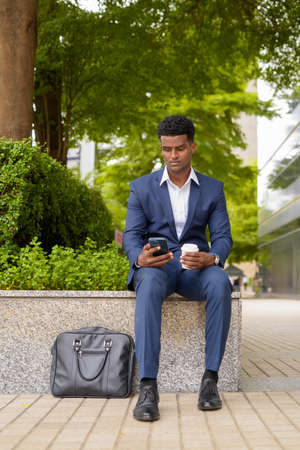 Portrait of African businessman using phone and holding take away coffee cup outdoors in city vertical shotの写真素材