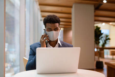 Portrait of African businessman wearing face mask and using laptop computer while talking on mobile phone in coffee shop horizontal shotの写真素材