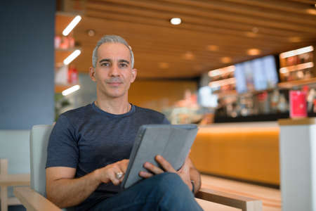 Portrait of handsome man sitting at coffee shop while holding digital tablet and looking at camera horizontal shot with copy spaceの写真素材