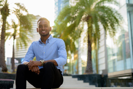 Portrait of handsome black African businessman sitting outdoors in city during summer
