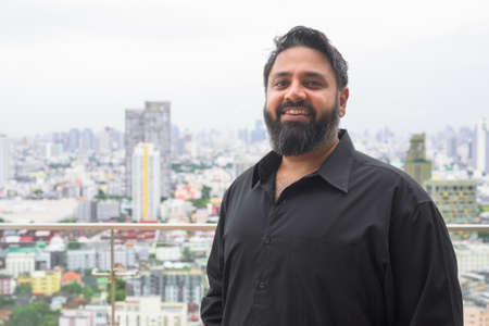 Horizontal portrait of handsome bearded Indian man smiling at rooftop in cityの写真素材