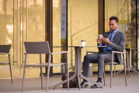 Handsome young African businessman wearing suit in city and sitting at coffee shop