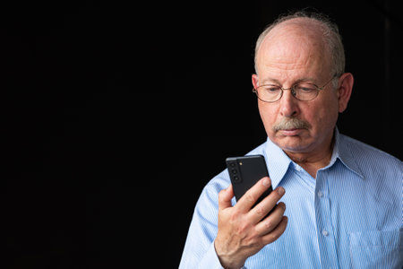 Grey haired senior man with mustache using mobile phone against black backgroundの写真素材