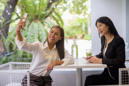 Two Asian women having a pleasant conversation over coffee in a modern cafe setting with greenery outside.の写真素材