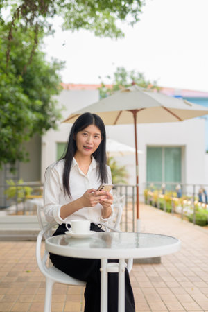 Professional Asian woman at an outdoor cafe with an umbrella using mobile phoneの写真素材