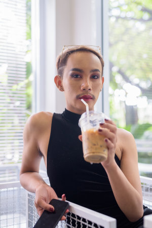 Portrait of beautiful Asian queer LGBT community supporter man with mustache wearing lipstick and jumpsuit in a bright, modern cafe with iced coffee and greeneryの写真素材