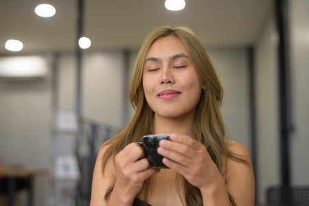 Portrait of beautiful young Asian woman sitting in cafe relaxing and happily holding coffee cup with eyes closed while thinking and daydreamingの写真素材