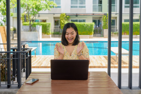 Asian woman outside with a swimming pool in the background. She is dressed in a yellow floral dress and sitting while using laptop computer while looking excited like she got some exciting good news.の写真素材