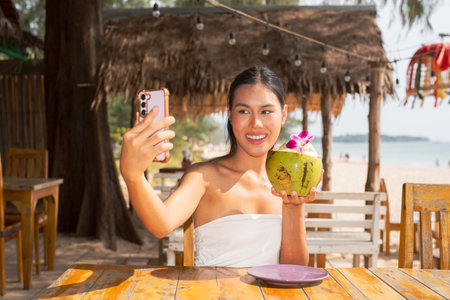 Happy young Asian woman relaxing on the beach holding fresh coconut juice while taking selfie in islandの写真素材