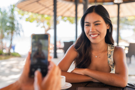 Young Asian woman smiling while friend taking photo of her with mobile phoneの写真素材