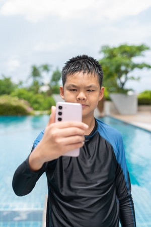 Asian teenage boy using phone after swimming in pool during summerの写真素材