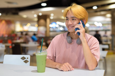 Thai gay teenager man with headphones using phone and drinking iced latte green tea in modern cafeの写真素材