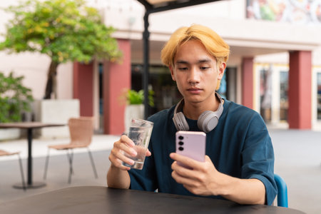 Asian gay teenager drinking water and using phone outdoors in cafe restaurant during summer