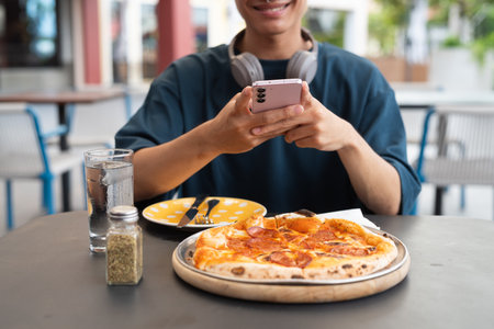 Young man taking photo of pepperoni pizza in outdoor restaurant with phoneの写真素材
