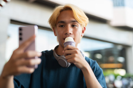 Young teen man eating soft serve ice cream outdoors and using smartphoneの写真素材