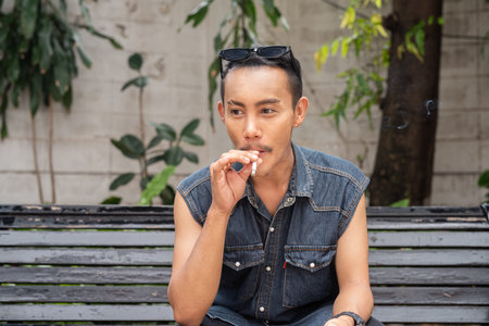 Stylish gay man sitting outdoors and smoking cigarette during summer