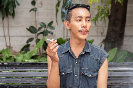 Stylish gay man sitting outdoors and smoking cigarette during summerの写真素材