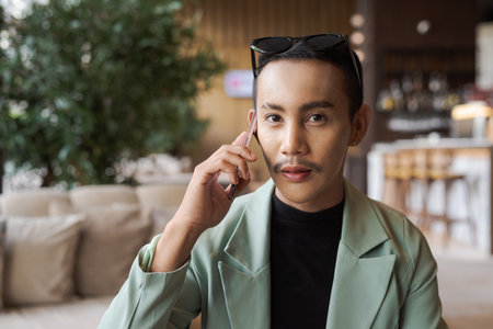Portrait of nonbinary genderfluid young Thai gay businessman using phone in cafeの写真素材