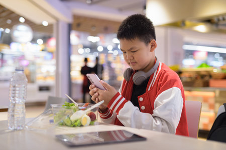 Young Teen Thai boy wearing headphones eating salad in cafeteria with phoneの写真素材