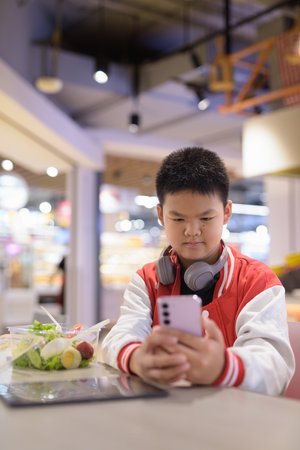 Young Teen Thai boy wearing headphones eating salad in cafeteria with phoneの写真素材