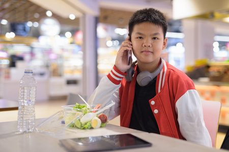 Young Teen Thai boy wearing headphones eating salad in cafeteria with phoneの写真素材