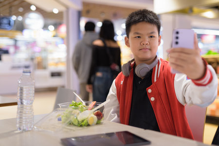 Young Teen Thai boy wearing headphones eating salad in cafeteria with phoneの写真素材