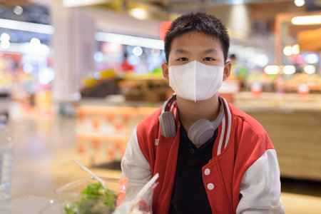 Thai teenage boy wearing face mask and headphones ready to eat salad indoorsの写真素材