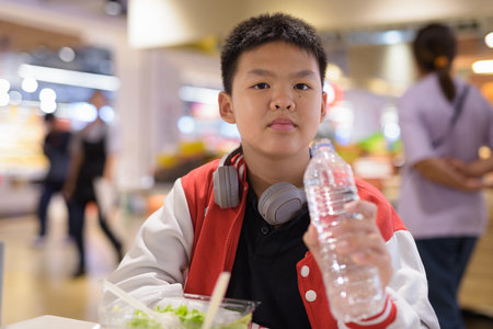 Thai teenage boy eating salad and drinking water indoors in cafe aloneの写真素材