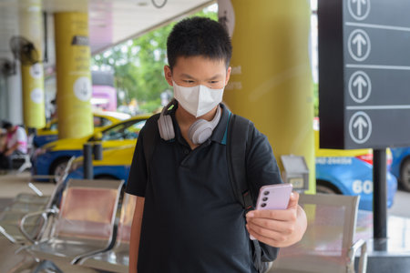 Masked Thai boy using phone with taxis in background wearing headphonesの写真素材