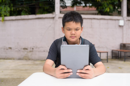 Thai teenage boy sitting at outdoor table using tablet wearing headphones during summer