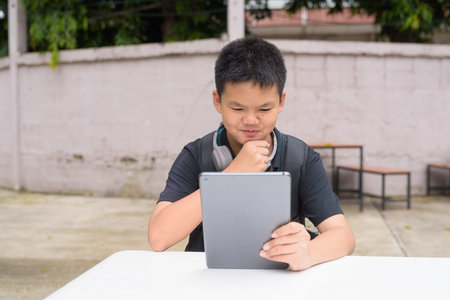 Thai teenage boy sitting at outdoor table using tablet wearing headphones during summer