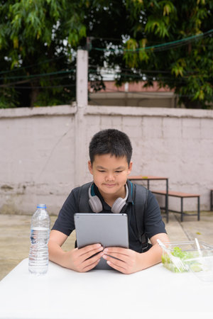 Thai student boy eating salad and using wireless technology at outdoor tableの写真素材