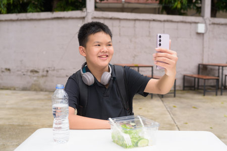 Thai student boy eating salad and using wireless technology at outdoor tableの写真素材