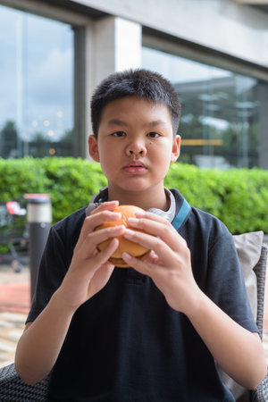 Thai student boy wearing headphones eating burger at outdoor table