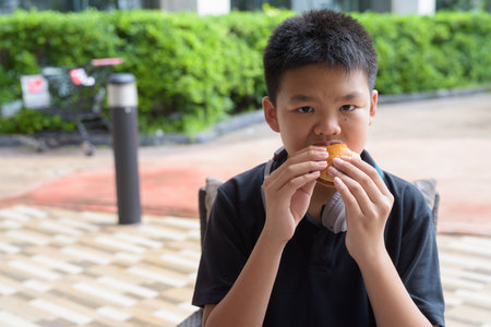 Thai student boy wearing headphones eating burger at outdoor tableの写真素材