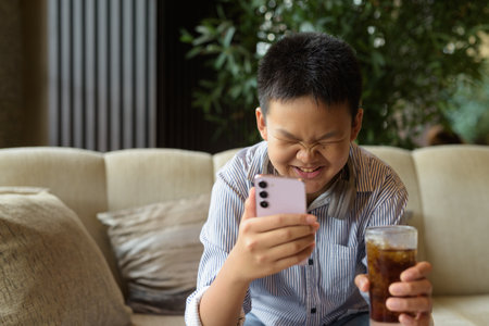 Thai boy drinking soda and using phone at restaurant with headphonesの写真素材