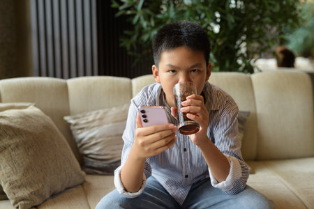 Thai boy drinking soda and using phone at restaurant with headphonesの写真素材