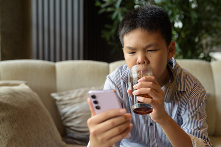 Thai boy drinking soda and using phone at restaurant with headphonesの写真素材