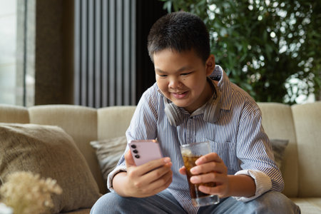 Thai boy drinking soda and using phone at restaurant with headphonesの写真素材