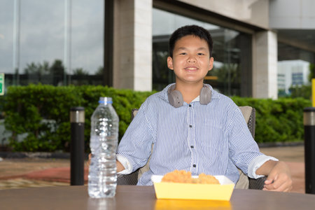 Thai student boy eating fried chicken with headphones at outdoor tableの写真素材