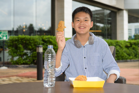 Thai student boy eating fried chicken with headphones at outdoor tableの写真素材