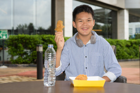 Thai student boy eating fried chicken with headphones at outdoor tableの写真素材