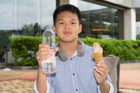 Thai student boy eating fried chicken and drinking water outdoorsの写真素材